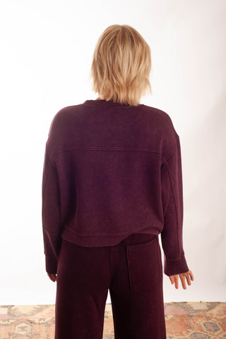A model viewed from behind wearing a dark plum purple cropped long-sleeve sweatshirt and matching wide-leg pants against a white studio background.