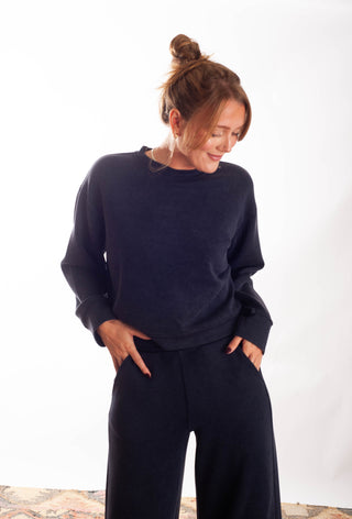 A woman wearing a dark navy blue crewneck sweatshirt and matching wide-leg sweatpants, standing against a plain white studio background.