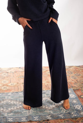 A person wearing dark navy wide-leg pants and a matching dark top, standing barefoot on a patterned rug against a plain white wall.
