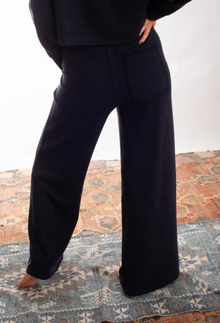 A person wearing dark navy wide-leg pants and a matching dark top, standing barefoot on a patterned rug against a white studio background.