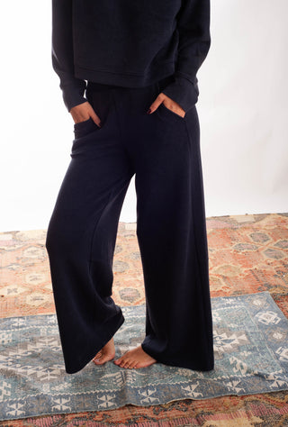 A person wearing dark navy wide-leg pants and a matching dark top, standing barefoot on a patterned rug against a plain white wall.