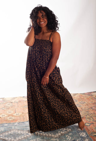 A woman modeling a sleeveless leopard-print maxi dress with thin straps, standing barefoot on a patterned rug against a white studio backdrop.