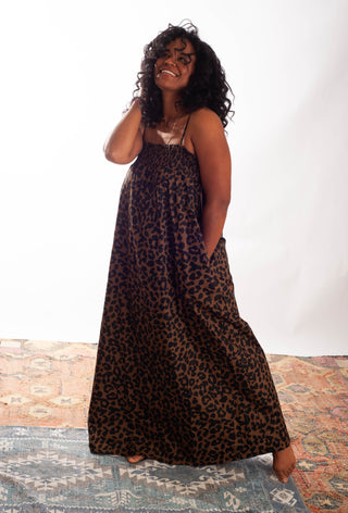 A woman modeling a sleeveless leopard-print maxi dress with thin straps and side pockets, standing barefoot on a patterned rug against a white studio backdrop.