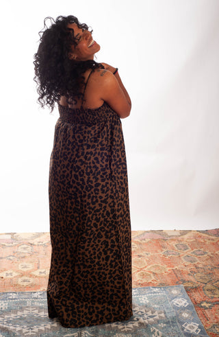 A woman with curly black hair is shown from the back wearing a leopard print maxi dress. She is standing on a patterned rug against a white studio backdrop.