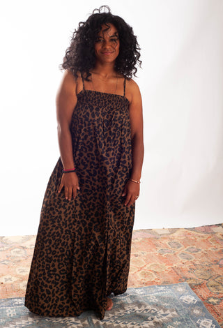 A woman with curly black hair wearing a leopard-print maxi dress with thin spaghetti straps, standing against a white studio backdrop.