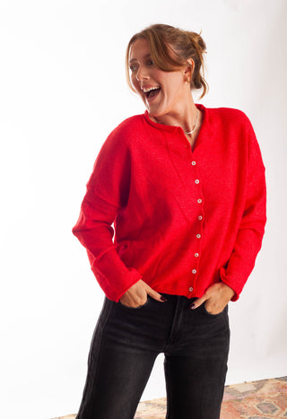 A woman wearing a bright red knit cardigan sweater with a button-down front and black jeans, standing against a plain white studio background.