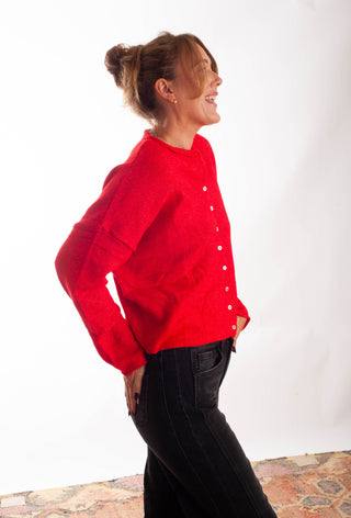 A woman wearing a bright red knit cardigan with white buttons and black jeans, standing in profile against a white studio background.