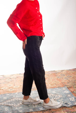 A person wearing a bright red long-sleeve button-front top, black jeans, and white low-top sneakers, standing on a patterned rug against a plain white studio background.