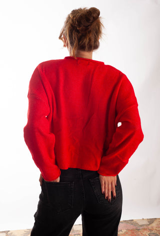 A person viewed from behind wearing a bright red knit sweater and black jeans against a plain white studio background.