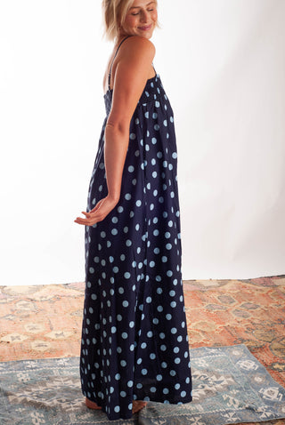 A woman modeling a navy blue maxi dress with a light blue polka dot pattern, standing on a patterned rug against a white studio background.