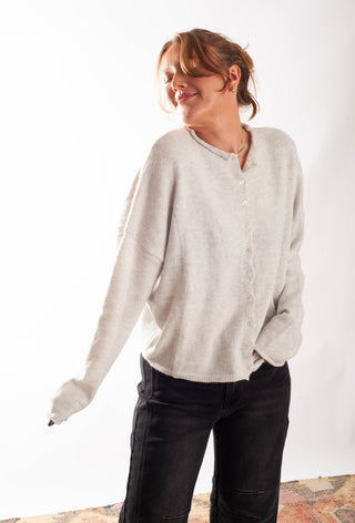 A woman wearing a light beige knit button-front cardigan sweater and black jeans, standing against a plain white studio background.