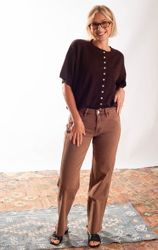 A woman modeling a dark brown short-sleeve button-front top and brown pants, standing on a patterned rug against a white studio background.