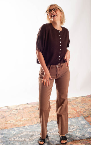A woman wearing a dark brown short-sleeve button-front top and brown pants, standing on a patterned rug against a white studio background.