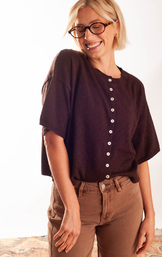 A woman wearing a dark brown short-sleeve button-up top and brown pants, smiling and looking down. She is wearing glasses and standing against a plain white background.