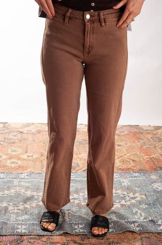 A person wearing brown straight-leg jeans, standing on a patterned rug with a white background behind.