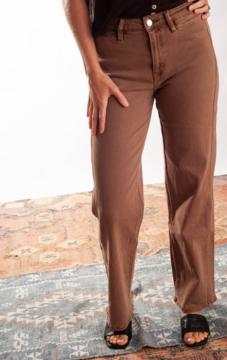 A person wearing brown straight-leg jeans with a black top and black slide sandals, standing on a patterned rug against a white studio background.