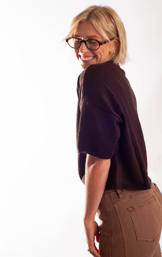 A woman wearing a dark brown short-sleeve button-up top and brown pants, smiling and looking down. She is wearing glasses and standing against a plain white background.
