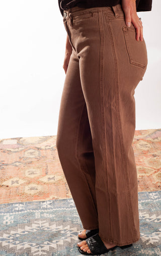 A person wearing brown wide-leg cropped pants with vertical seam detailing, paired with black slide sandals, standing on a patterned rug against a white wall.