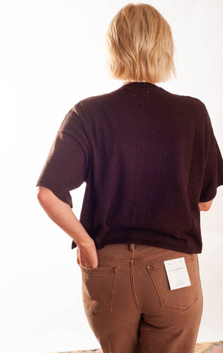 A woman wearing a dark brown short-sleeve button-up top and brown pants, smiling and looking down. She is wearing glasses and standing against a plain white background.