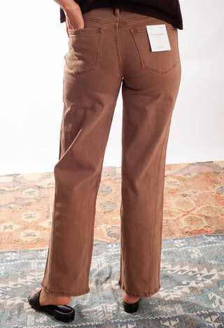A person shown from the back wearing brown wide-leg jeans, standing on a patterned rug. The jeans have a white paper tag attached to the back pocket.