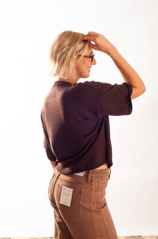 A woman with short blonde hair and glasses, shown from a side-back angle, wearing a dark brown textured short-sleeve top and brown pants against a plain white background.