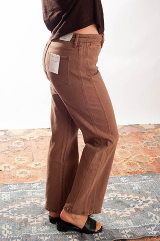 A person wearing brown wide-leg jeans with a side pocket and a white paper tag attached near the back waistband, standing on a patterned rug against a white wall.