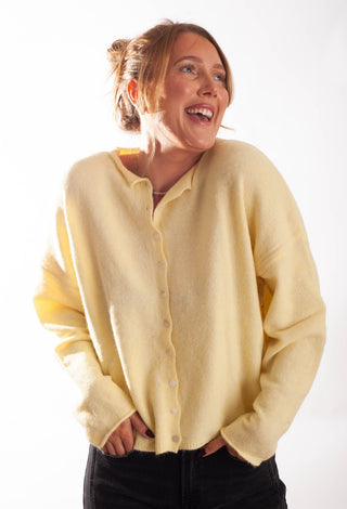 A woman wearing a soft, pale yellow button-up cardigan sweater and black pants, smiling and looking off-camera against a bright white background.