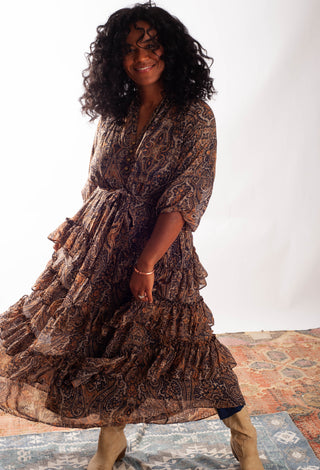 A woman wearing a brown and black paisley print tiered midi dress with a tie waist, standing on a patterned rug against a white studio background.