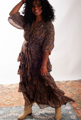 A woman modeling a brown and black paisley print maxi dress with tiered ruffles and elbow-length sleeves, paired with beige ankle boots, standing on a patterned rug against a white studio background.