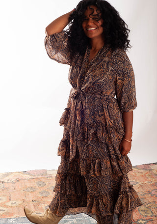 A woman wearing a brown and black paisley print tiered midi dress with a tie waist, standing on a patterned rug against a white studio background.