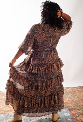 A woman viewed from behind wearing a brown and black paisley tiered maxi dress, holding up the side of the dress to show the layers and fabric. She is standing on a patterned rug against a white studio background.