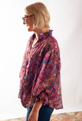 A woman with short blonde hair and glasses, shown in profile wearing a loose, long-sleeve button-up blouse with a purple, pink, and orange floral pattern, paired with blue jeans against a plain white background.