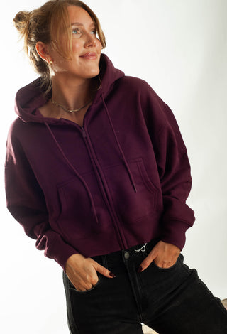 A woman wearing a cropped, zip-up hoodie with black jeans, standing against a plain white studio background.
