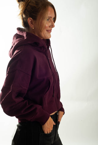 A woman wearing a cropped, zip-up hoodie with black jeans, standing against a plain white studio background.