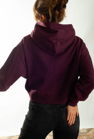 A woman wearing a cropped, zip-up hoodie with black jeans, standing against a plain white studio background.