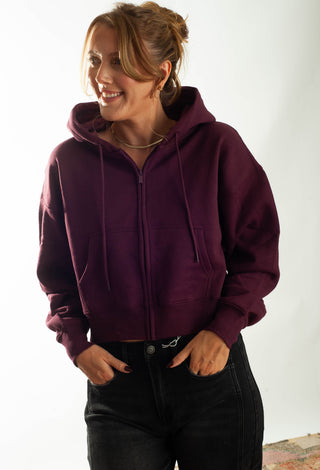 A woman wearing a cropped, zip-up hoodie in deep burgundy with black jeans, standing against a plain white studio background.