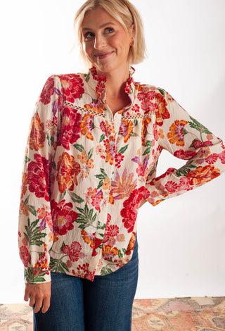 A woman wearing a long-sleeve floral blouse with a high gathered neckline and blue jeans, standing against a plain white background.