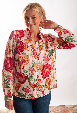 A woman wearing a long-sleeve button-up blouse with a vibrant red, orange, and green floral print on a cream background, paired with blue jeans, set against a plain white studio background.