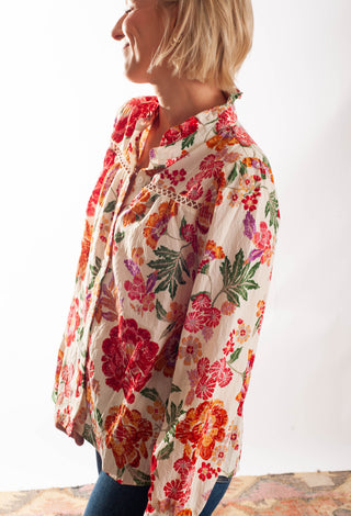 A woman wearing a long-sleeve bohemian-style button-up blouse with a vibrant multicolor floral print on a cream background, paired with blue jeans.