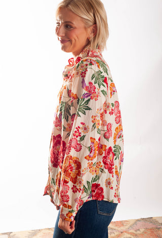 A woman wearing a long-sleeve button-up shirt with a vibrant multicolor floral print on a cream background, paired with blue jeans, photographed against a plain white studio background.