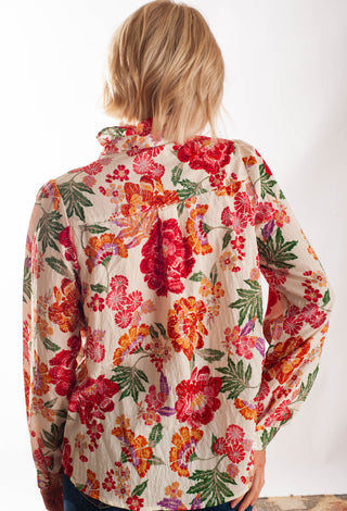 A woman viewed from behind wearing a long-sleeve button-up shirt with a vibrant, multicolor floral print on a textured cream fabric, set against a plain white background.