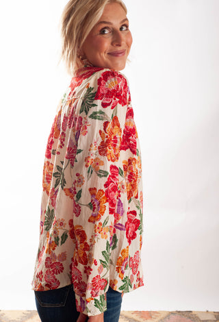 A woman wearing a cream-colored long-sleeve blouse with a vibrant red, orange, and green floral print, paired with dark blue jeans, photographed against a plain white background.