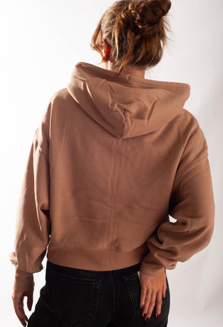 A person shown from the back wearing a cropped brown hoodie and black jeans against a plain white studio background.