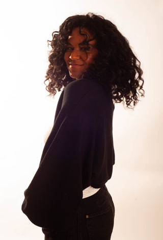 A woman with long, curly black hair is shown from the back and side, wearing a black oversized long-sleeve top and black pants against a bright white studio background.
