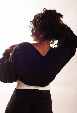 A person shown from the back wearing a black cropped V-neck sweater over a white top and black jeans, against a plain white studio background.