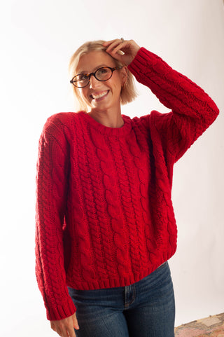 A smiling woman with short blonde hair and glasses wearing a bright red cable-knit sweater and blue jeans, posing against a plain white background.