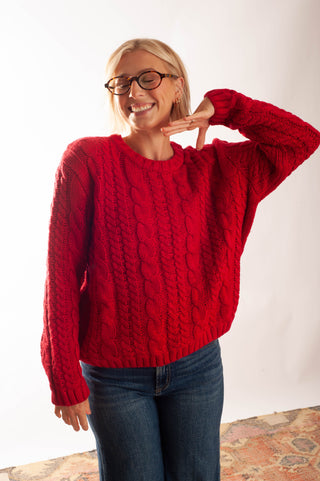 A smiling blonde woman wearing round black eyeglasses and a chunky red cable-knit sweater with blue jeans, posing against a white studio background.