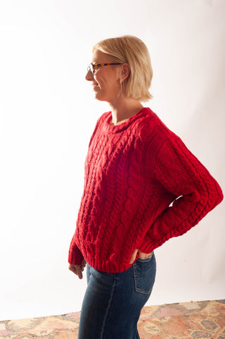 A woman with short blonde hair and glasses wearing a red cable-knit sweater and blue jeans, standing in profile against a white studio background.