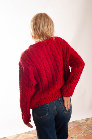 A smiling woman with short blonde hair and glasses wearing a bright red cable-knit sweater and blue jeans, posing against a plain white background.