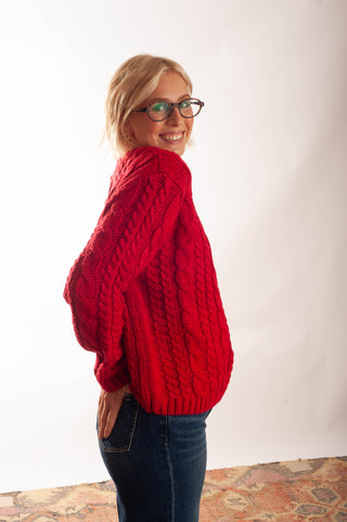 A woman with blonde hair and glasses, wearing a bright red cable-knit sweater and dark blue jeans, posing in a studio setting against a white background.